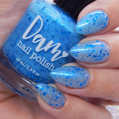 Blueberry Sanctuary by Dam Nail Polish, sheer light blue crelly with vivid blue hex glitters, blue-to-purple shifting shimmer, and holographic micro-glitter dusting, bottle held in a light hand