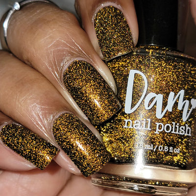 Champagne Sparkles by Dam Nail Polish, deep black-brown jelly with dense bronze shimmer and bright gold reflective glitter under indoor warm light, bottle held in a medium skin tone hand