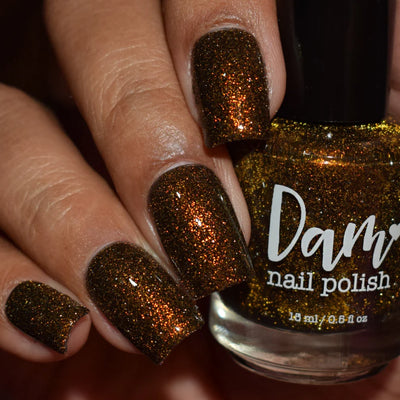 Champagne Sparkles by Dam Nail Polish, dark brown-black jelly with dense copper-bronze shimmer and gold reflective glitter under indoor warm light, bottle held in a light-medium skin tone hand