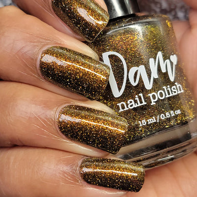 Champagne Sparkles by Dam Nail Polish, dark brown-black jelly with bronze-gold shimmer and sparkling gold reflective glitter under indoor warm light, bottle held in a light-medium skin tone hand