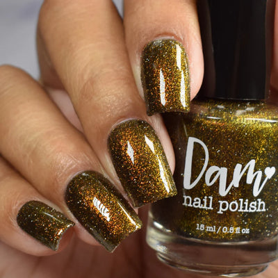 Champagne Sparkles by Dam Nail Polish, black-gold jelly with bronze shimmer and gold reflective glitter reading deep olive-bronze under studio light, bottle held in a light-medium skin tone hand