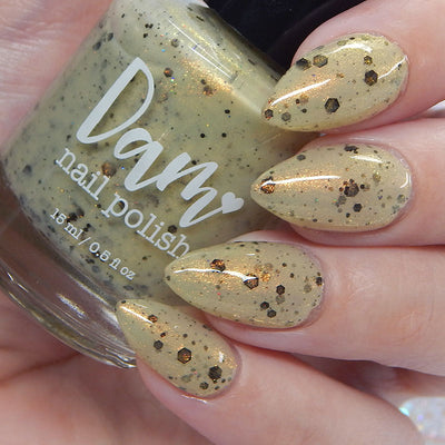 OG Devo-tea by Dam Nail Polish, warm tan crelly with gold shimmer and scattered bronze hex plus small black glitters, on a light skin tone hand