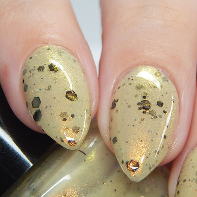 OG Devo-tea by Dam Nail Polish, warm tan crelly showing gold shimmer, black hex glitters, and sparkling copper-gold glitter flakes, on a light skin tone hand