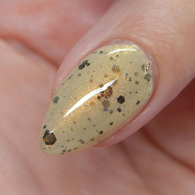 OG Devo-tea by Dam Nail Polish, tan crelly with dense gold shimmer and scattered black hex and micro glitters on a single nail, on a light skin tone hand