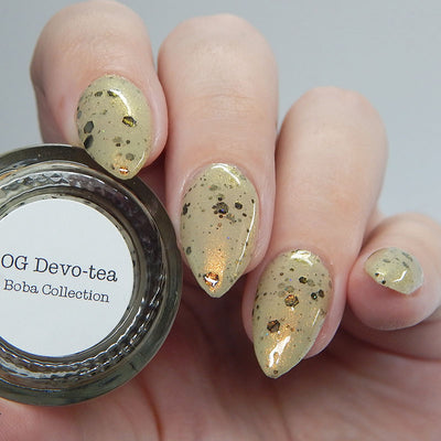 OG Devo-tea by Dam Nail Polish, warm tan-khaki crelly with gold shimmer and dark olive-black glitters of varying sizes, on a light skin tone hand