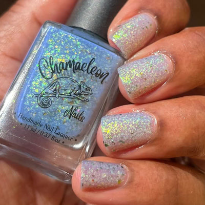The Forgotten Lagoon (UV Polish) by Chamaeleon Nails, flakie UV with sheer white-lavender base and periwinkle blue UV-activated bottle in sunlight, green, gold, and orange multichrome flakies, bottle held in a medium hand