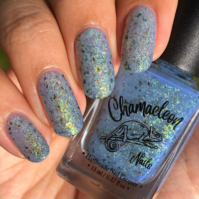 The Forgotten Lagoon (UV Polish) by Chamaeleon Nails, blue-grey flakie UV in UV-activated state with dense green, orange, and bronze multichrome flakes in bright sunlight, bottle held in a light-medium hand