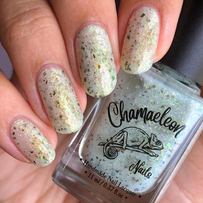 The Forgotten Lagoon (UV Polish) by Chamaeleon Nails, warm white-grey flakie UV in resting state with green, gold, and bronze iridescent and ultrachrome flakes under indoor warm light, bottle held in a light-medium hand
