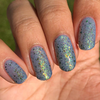 The Forgotten Lagoon (UV Polish) by Chamaeleon Nails, flakie UV in full UV-activated state with white base shifted to periwinkle blue, dense green-to-orange iridescent and bronze ultrachrome flakes in bright sunlight, on a light-medium skin tone hand