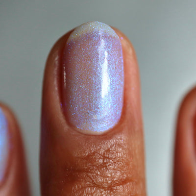 The Fire's Burning Blue by Cadillacquer, soft lavender-blue base with blue shimmer particles and pink-white highlight shift in studio light, on a medium-deep skin tone hand
