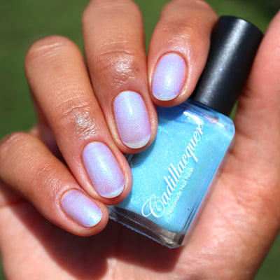 The Fire's Burning Blue by Cadillacquer, sky blue in bottle shifting to pearlescent lavender-blue with pink-to-purple shimmer on nails, bottle held in a light-medium hand
