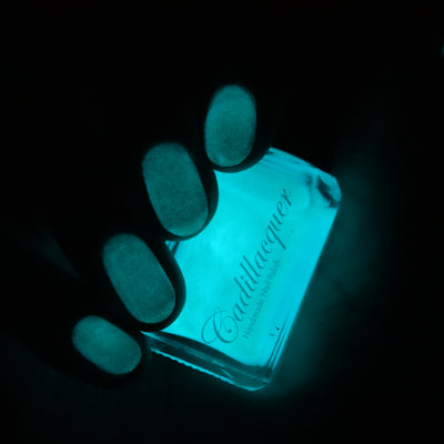 The Fire's Burning Blue by Cadillacquer, glow-in-the-dark activated state showing vivid cyan-teal luminescent glow, bottle standalone product shot