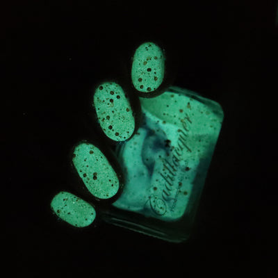 Light the Flame by Cadillacquer, mint-green glow-in-the-dark activated state with scattered dark reddish-brown metallic glitter, bottle held in hand