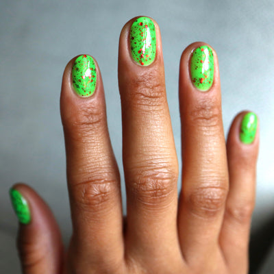 Light the Flame by Cadillacquer, neon green creme glitter glow-in-the-dark with dispersed red metallic glitter in bright daylight, on a medium-deep skin tone hand
