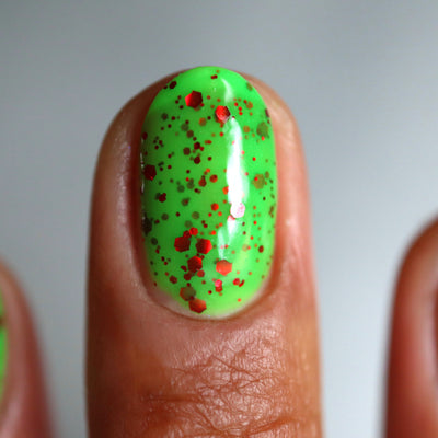 Light the Flame by Cadillacquer, neon green glitter glow-in-the-dark with red metallic hex glitter in micro-to-medium sizes, on a medium-deep skin tone hand