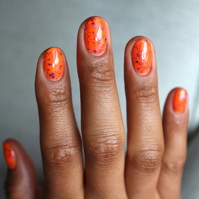 Endure And Survive by Cadillacquer, neon orange glitter with metallic purple and dark violet micro specks across five nails, on a medium-deep skin tone hand