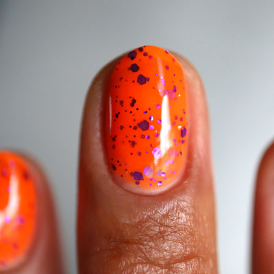 Endure And Survive by Cadillacquer, neon orange base with deep purple and bright magenta metallic glitter ranging from fine dots to hex shards, on a medium-deep skin tone hand