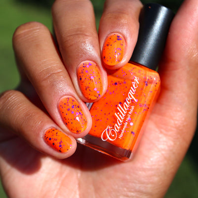 Endure And Survive by Cadillacquer, neon orange glitter with scattered metallic purple and magenta-pink particles in bright daylight, bottle held in a light-medium hand