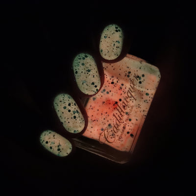 Endure And Survive by Cadillacquer, glow-in-the-dark activated state, nails emitting soft mint-green luminescence with dark speckled glitter, bottle glowing vivid coral-orange in darkness, bottle held in hand