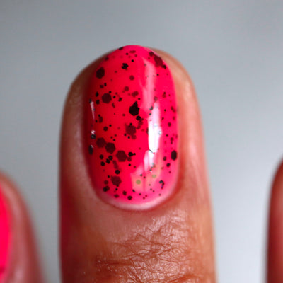 The Night's a Brighter Shade by Cadillacquer, vivid neon pink with speckled matte black glitter particles in studio light, on a medium-deep skin tone hand