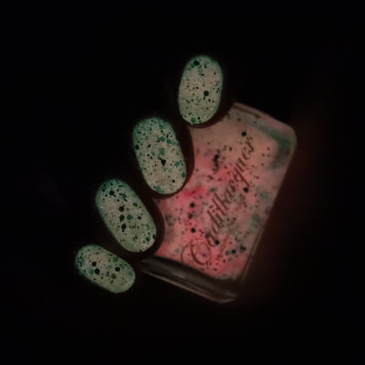 The Night's a Brighter Shade by Cadillacquer, coral-to-mint glow-in-the-dark activated state with matte black glitter particles, on a dark-lit swatch on nails