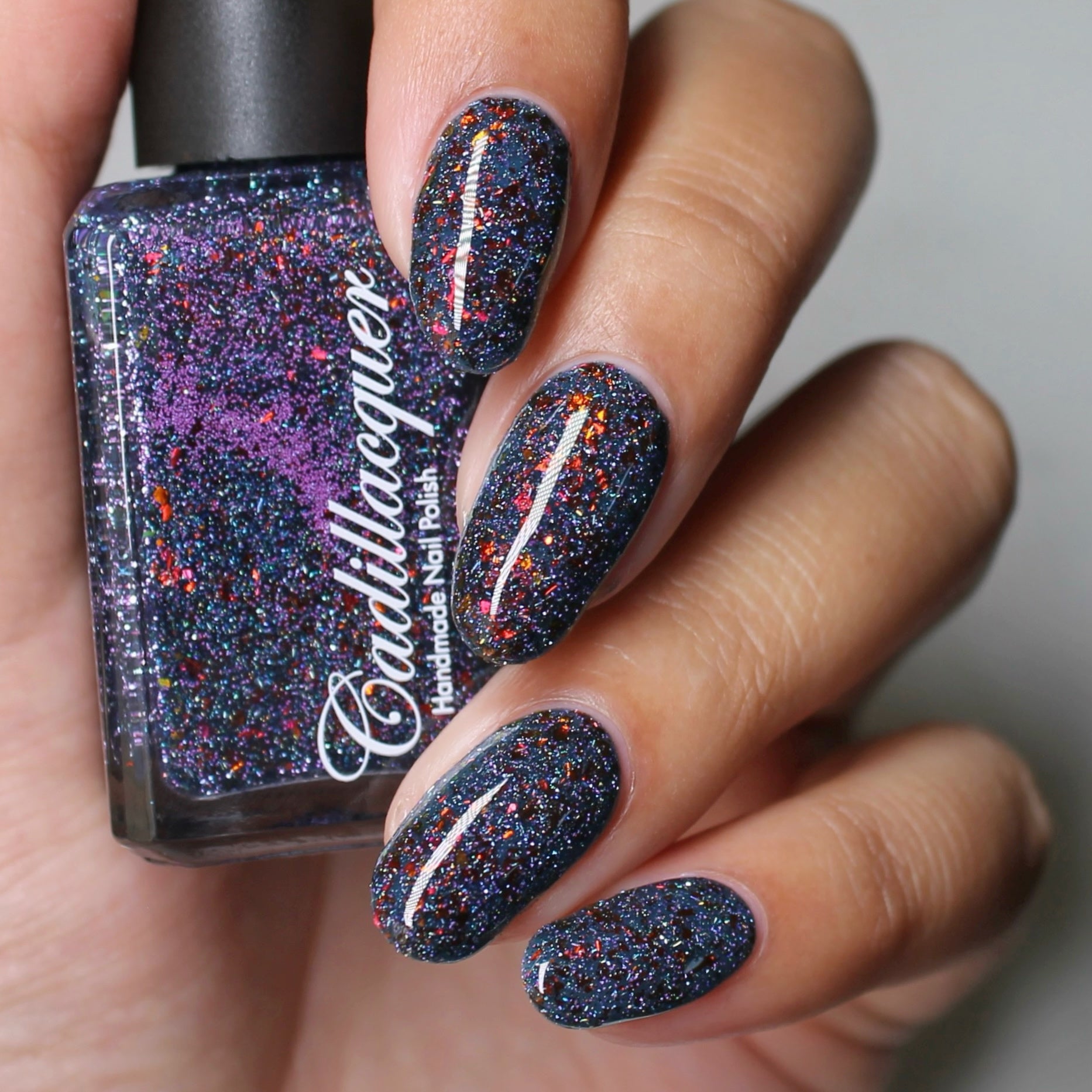 A bottle of Cadillacquer nail polish in a deep teal color with red/copper/gold chrome chameleon flakies and purple reflective glitter, shown alongside a swatch of the polish color.