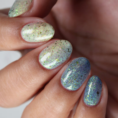 The Forgotten Lagoon (UV Polish) by Chamaeleon Nails, flakie UV showing photochromic range, sheer milky white on index to UV-activated blue on ring and pinky, with green-gold iridescent and ultrachrome flakes, on a light-medium skin tone hand