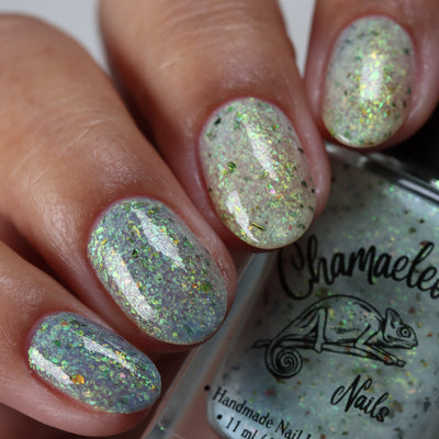 The Forgotten Lagoon (UV Polish) by Chamaeleon Nails, sheer milky white flakie UV base in resting state with large green-to-orange multichrome and bronze UCC flakes, on a light-medium skin tone hand
