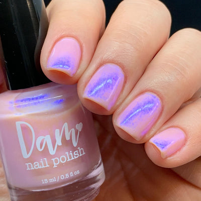 A Whimsical Dream by Dam Nail Polish, sheer blush pink jelly with vivid blurple shimmer flashing at center-nail under bright daylight, bottle held in a light skin tone hand