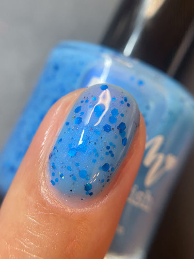 Blueberry Sanctuary by Dam Nail Polish, sheer periwinkle blue crelly with assorted metallic blue hex glitters and blue-to-purple shimmer shift in warm indoor light, on a light skin tone hand