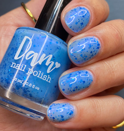 Blueberry Sanctuary by Dam Nail Polish, sheer cornflower blue crelly with metallic royal blue hex glitters and blue-violet shifting shimmer, bottle held in a light-medium hand