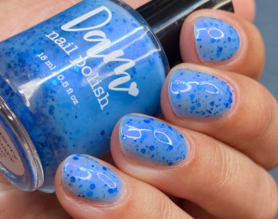 Blueberry Sanctuary by Dam Nail Polish, sheer light blue jelly crelly with assorted metallic blue glitters and holographic glitter dusting in bright daylight, bottle held in a light-medium hand