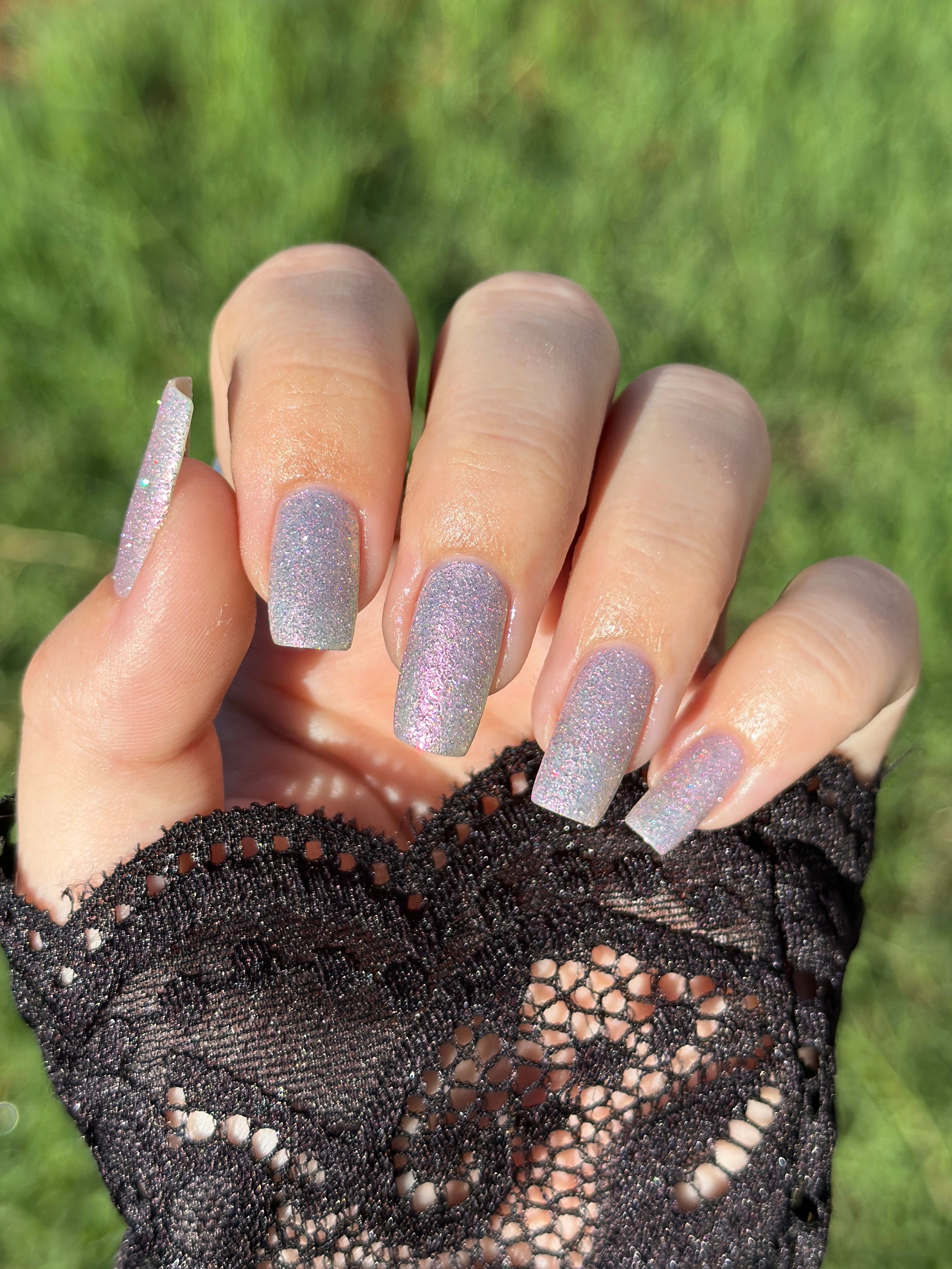 Lavender Haze by Glitch Lacquer bottle held in hand with one long almond nail swatched, very light purple polish with pink-yellow-green-teal multichrome reflect and red reflective glitters, medium-deep skin tone, studio lighting.