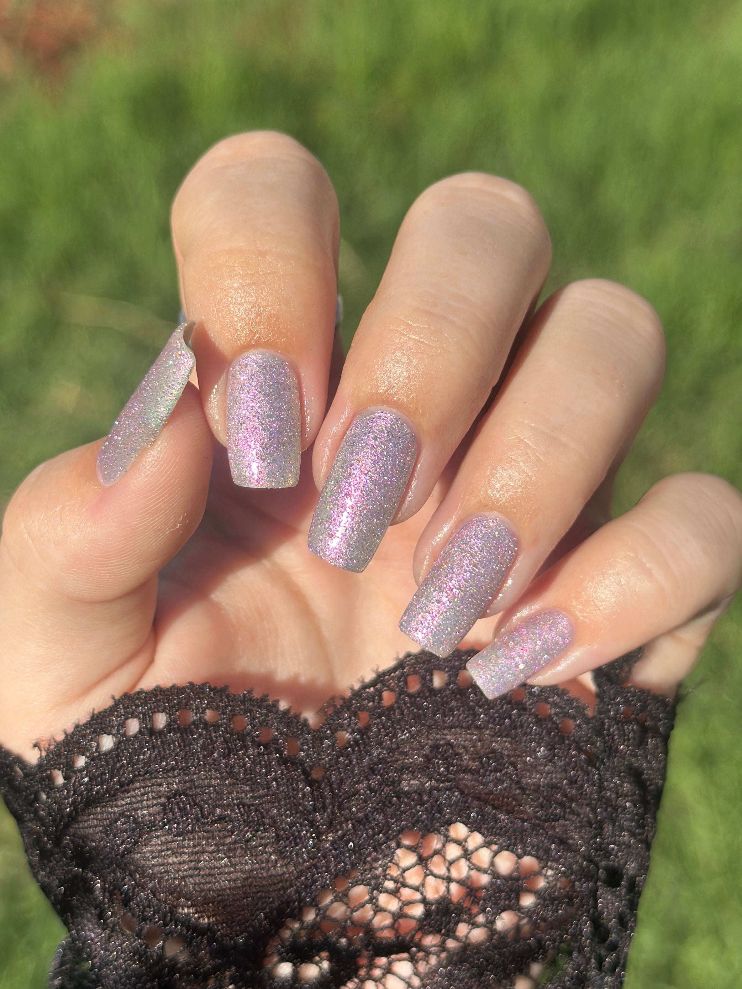 Lavender Haze by Glitch Lacquer bottle held in hand with one long coffin nail swatched outdoors, very light purple polish with pink-yellow-green-teal multichrome reflect and red reflective glitters, light skin tone, bright daylight.