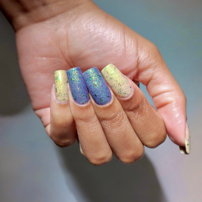 The Forgotten Lagoon (UV Polish) by Chamaeleon Nails, flakie UV on five coffin nails showing dual-tone UV reaction, cream-white resting state on index and pinky beside periwinkle-blue activated middle and ring nails, with green iridescent and bronze UCC flakes, on a medium-deep skin tone hand