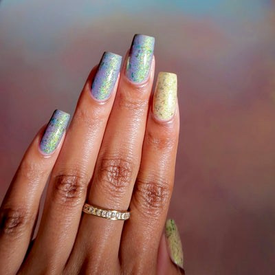 The Forgotten Lagoon (UV Polish) by Chamaeleon Nails, flakie UV on five long square nails, mostly soft lavender-blue UV-activated with dense green and orange iridescent flakes, cream-white resting state visible on index finger, on a medium-deep skin tone hand