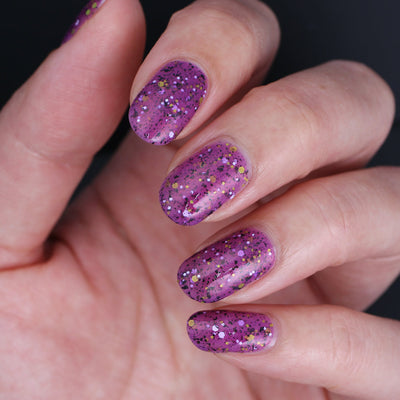 AHHHH The Flakies by Dam Nail Polish, vivid magenta-purple jelly with black flakies and gold, white, and light purple hex glitters, on a light skin tone hand