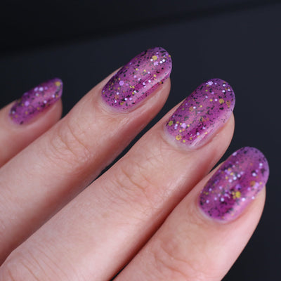 AHHHH The Flakies by Dam Nail Polish, purple-magenta shimmer glitter jelly scattered with black flakies and white, yellow hex glitters, on a light skin tone hand