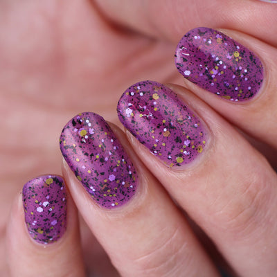AHHHH The Flakies by Dam Nail Polish, medium-deep purple jelly with black flakies, gold and white hex glitter particles, warm red-orange shimmer undertone, on a light skin tone hand