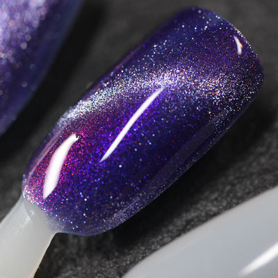 Grand Celebration by Dam Nail Polish, deep indigo-purple shimmer magnetic showing purple-to-fuchsia-to-silver multichrome shift with silver cat-eye streak, swatch stick