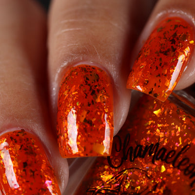 Sauron's Eye by Chamaeleon Nails, fiery orange jelly with gold-amber and copper-red multichrome flakies catching warm tones, on a light-medium skin tone hand