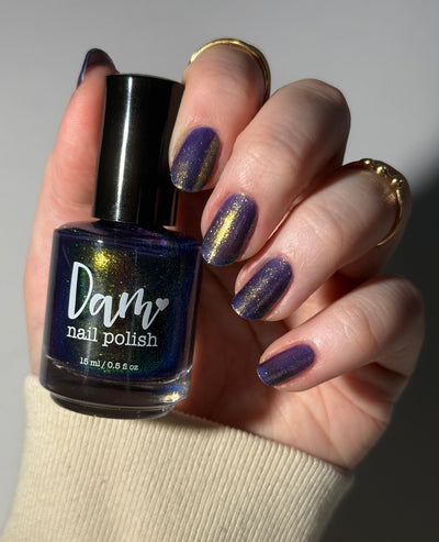 Sandman by Dam Nail Polish, cool blurple jelly with striking linear gold-to-green-to-blue multichrome shimmer shift, bottle held in a light skin tone hand