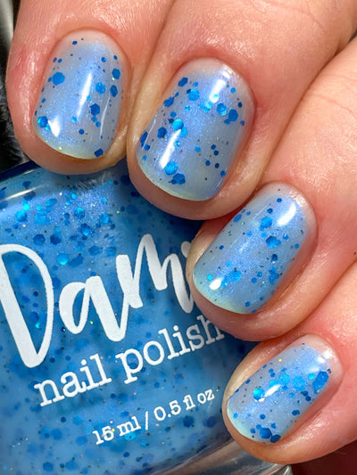 Blueberry Sanctuary by Dam Nail Polish, sheer light blue crelly with metallic blue hex glitters and soft blue-shifting iridescent shimmer in bright daylight, on a light skin tone hand