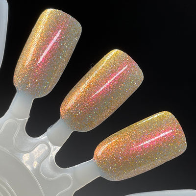 I Love Oklahoma by Dam Nail Polish, warm gold base with red-to-pink multichrome shimmer and scattered holographic micro glitter under direct flash, swatch stick