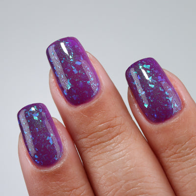 Purple Moon (UV Polish) by Chamaeleon Nails, deep violet-purple UV-activated state with teal, turquoise, and blue multichrome flakies and green shimmer, on a light-medium skin tone hand