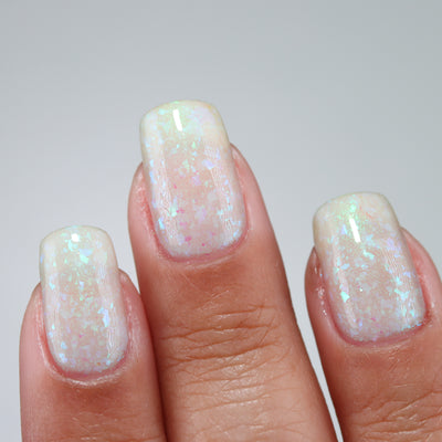 Purple Moon (UV Polish) by Chamaeleon Nails, opalescent creamy white resting state with iridescent blue-green and pink-shifting flakie particles, on a light-medium skin tone hand