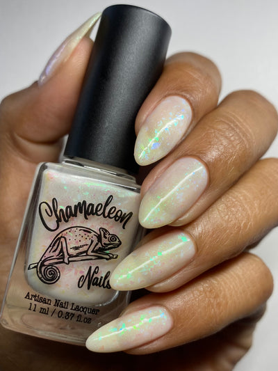 Purple Moon (UV Polish) by Chamaeleon Nails, creamy off-white resting state with blue-green-turquoise shifting flakies and shimmer, UV purple not activated under studio lighting, bottle held in a medium-deep hand