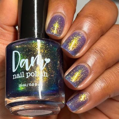 Sandman by Dam Nail Polish, cool blurple jelly with scattered gold and green shimmer sparkle, bottle held in a medium-deep skin tone hand