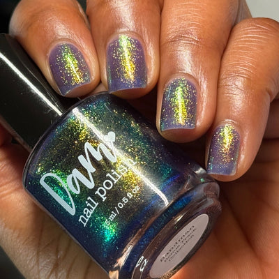Sandman by Dam Nail Polish, blurple jelly with gold-to-green-to-blue multichrome shimmer under warm indoor light, bottle held in a medium-deep skin tone hand