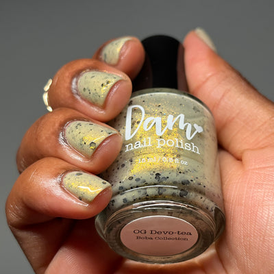OG Devo-tea by Dam Nail Polish, pale sage-tan crelly with gold shimmer and scattered black hex glitters, bottle held in a medium-deep skin tone hand
